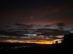 A breathtaking sunset over a mountainous landscape, featuring deep orange and yellow hues blending into darker blues and purples of the sky. Fluffy clouds are scattered throughout, illuminated by the fading daylight. A faint crescent moon can be seen above the horizon, adding a serene touch to the twilight scene. The silhouette of trees is visible in the foreground, while a city lights up in the valley below, contrasting with the natural beauty of the setting.