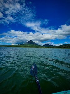 Kayaking on a lake with a paddle in the foreground, facing a volcano surrounded by greenery under a bright blue sky with scattered clouds