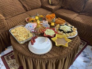 A cozy table with snacks like noodles, sauce, and sweets in small bowls. Colorful bangles, marigold flowers, and lit lamps create a festive feel, with a brown couch and decorative fan adding warmth.
