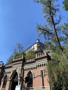 Low-angle view of a historic brick and stone building with arched windows and decorative battlements, trees on one side, a Mexican flag on top, and signs in front under a clear blue sky.