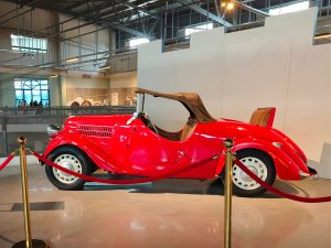 A bright red vintage convertible car with a brown soft top is displayed indoors, roped off with red ropes and gold stanchions.