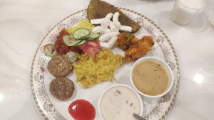 A plate of assorted Indian dishes with patties, vegetables, flatbread, sauces, and yogurt, served with a drink in the background.
