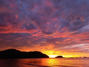 Sunset over the sea with a sky filled with clouds in shades of violet, pink, and deep orange. The sun sets on the horizon between two dark silhouetted islands, while several boats rest on calm waters reflecting golden and orange highlights.
