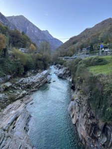 A calm Verzasca River flows through a narrow rocky gorge, surrounded by green and autumn mountains in Lavertezzo, Switzerland.