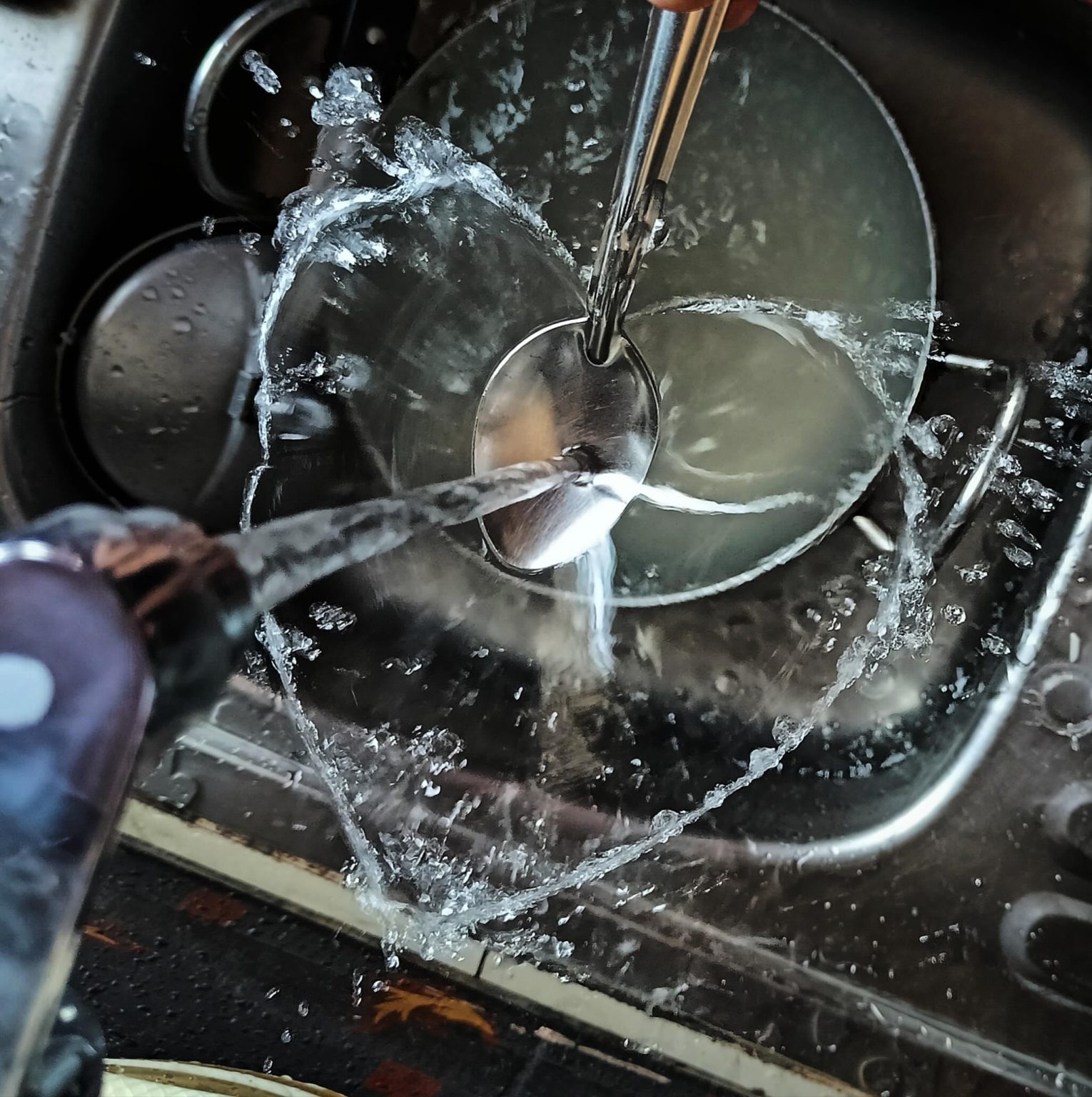 Water from a running tap splashes off a spoon in a steel kitchen sink, spray arcing across the bowl.