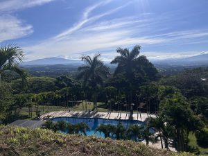 A scenic view of a swimming pool surrounded by lush greenery, with several tall palm trees lining the pool's edge. In the background, mountains are visible under a clear blue sky, with wispy clouds scattering across the horizon. The landscape below is dotted with trees and gently rolling hills, suggesting a tranquil and picturesque setting.