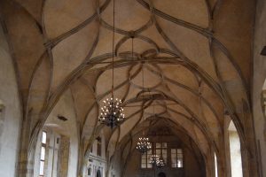 Interior view of a grand Gothic hall with intricate vaulted stone ceilings and ornate hanging chandeliers. A historical European hall featuring elegant arched architecture and warm lighting.