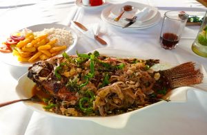 A whole cooked fish topped with onions and green pepper served on a white table with rice and chips.
