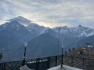 Panoramic mountain view with snow-capped peaks, huts, and a railing with a lantern under a cloudy sky.
