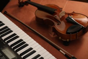 A viola and its bow rest on a surface beside a digital keyboard, showing the instrument’s wooden body, strings, and chin rest next to the black and white keys.