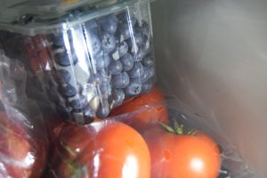 Fresh blueberries in a plastic container stacked on top of tomatoes in a refrigerator.