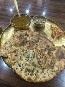 Golden plate with two spiced kulchas, a small bowl of chole (chickpeas), a side of chutney, and sliced onions on a wooden table.