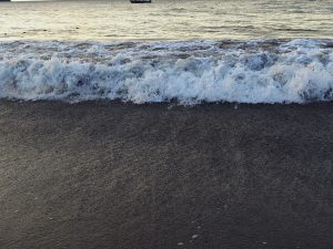 Dark, coarse sand meets foamy white waves crashing onto shore, with calm ocean extending to horizon under soft lighting.