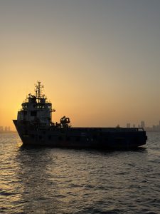 A tugboat/supply ship in harbor off the coast of India. It's silhouetted against the setting sun, and you can see a city skyline in the distance.