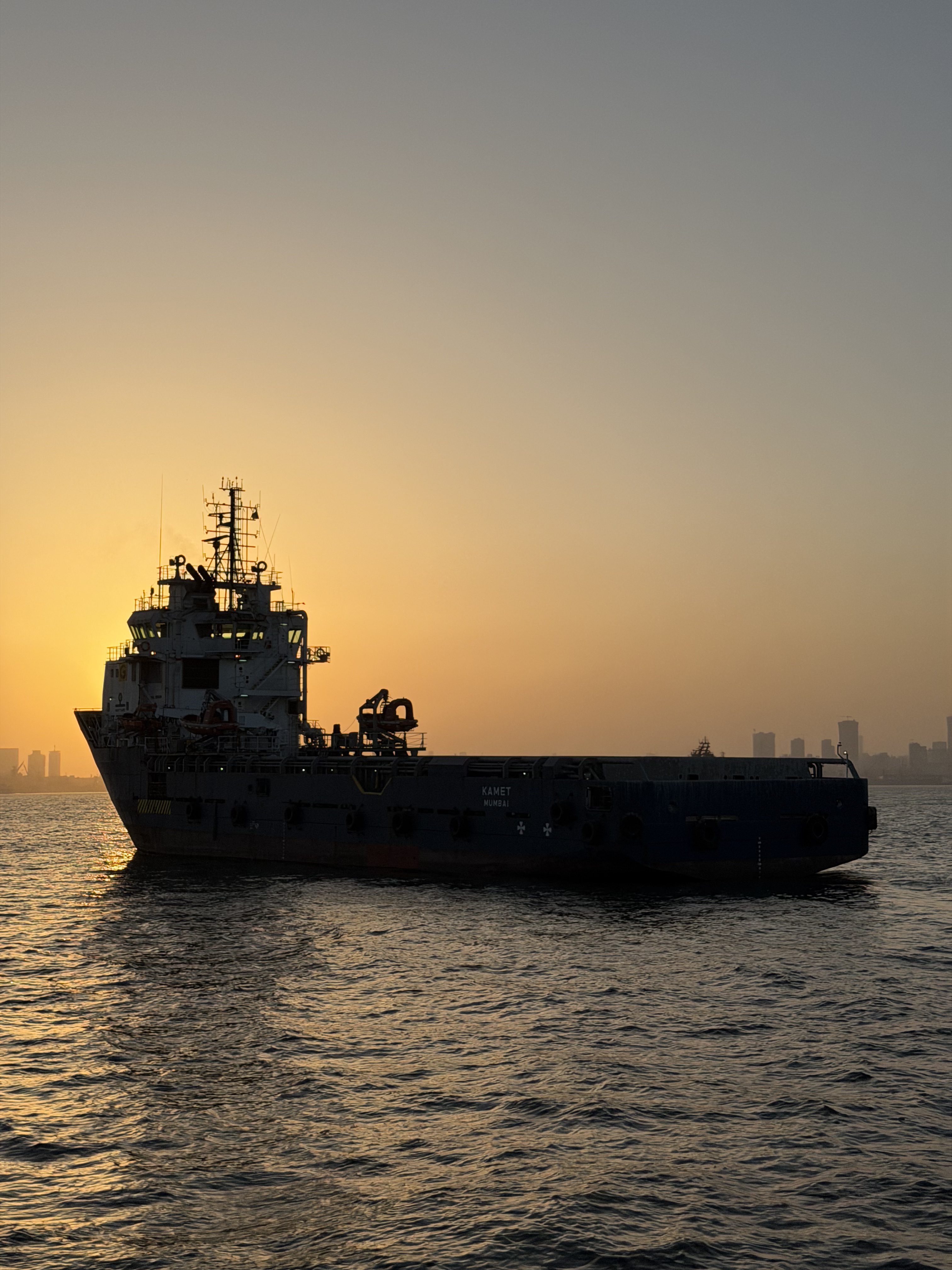 A tugboat/supply ship in harbor off the coast of India. It's silhouetted against the setting sun, and you can see a city skyline in the distance.