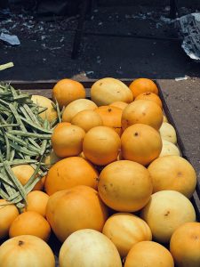 A heap of round yellow melons is displayed on a wooden surface, with green vegetables nearby.
