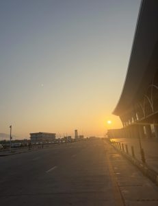 Sunset scene at an airport, showing an empty road leading to a terminal. The sun sets on the horizon, casting a warm glow and creating a serene atmosphere.