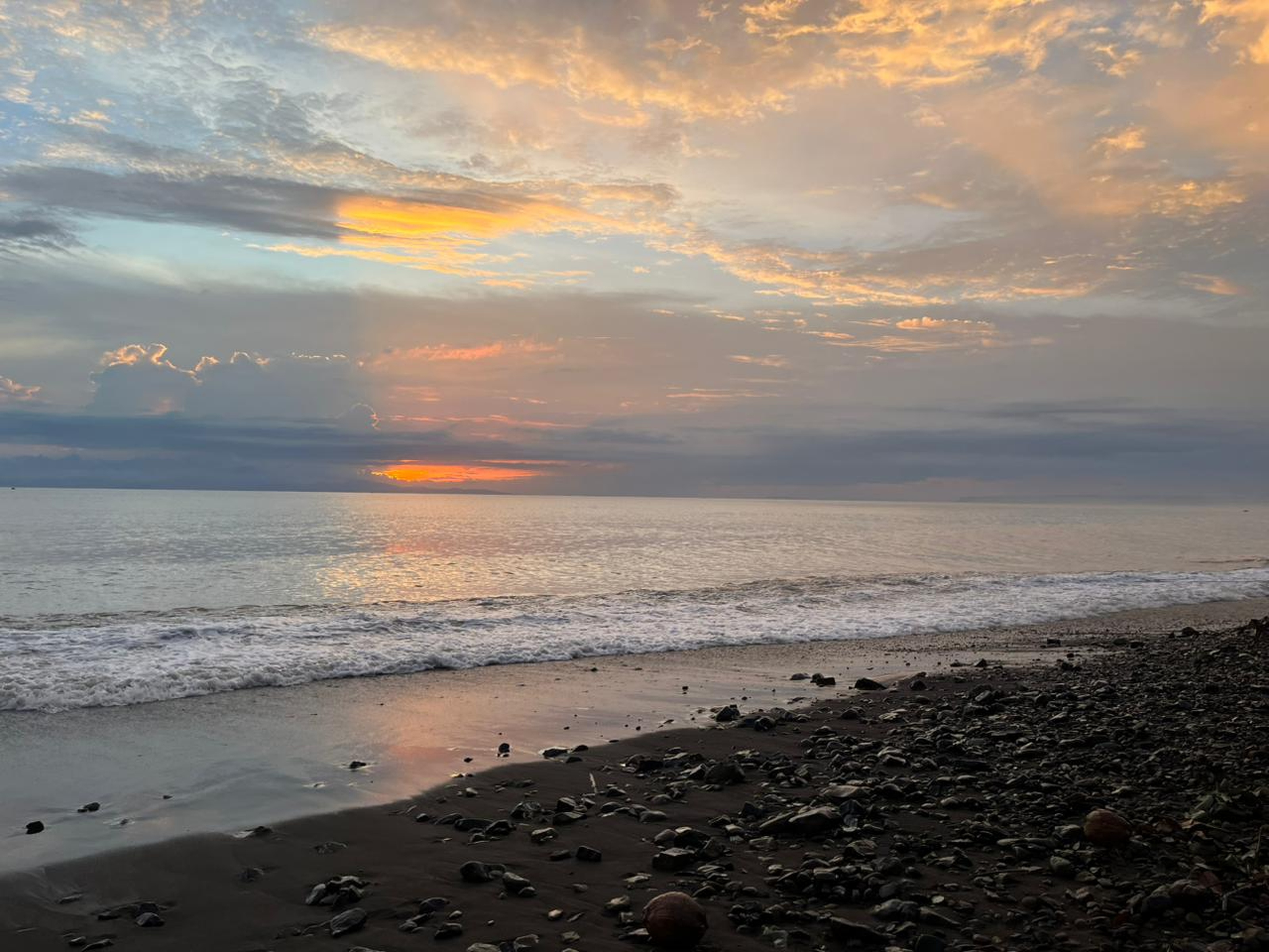 A wide ocean beach at sunset with golden and orange clouds reflected on the wet sand and rocky shore
