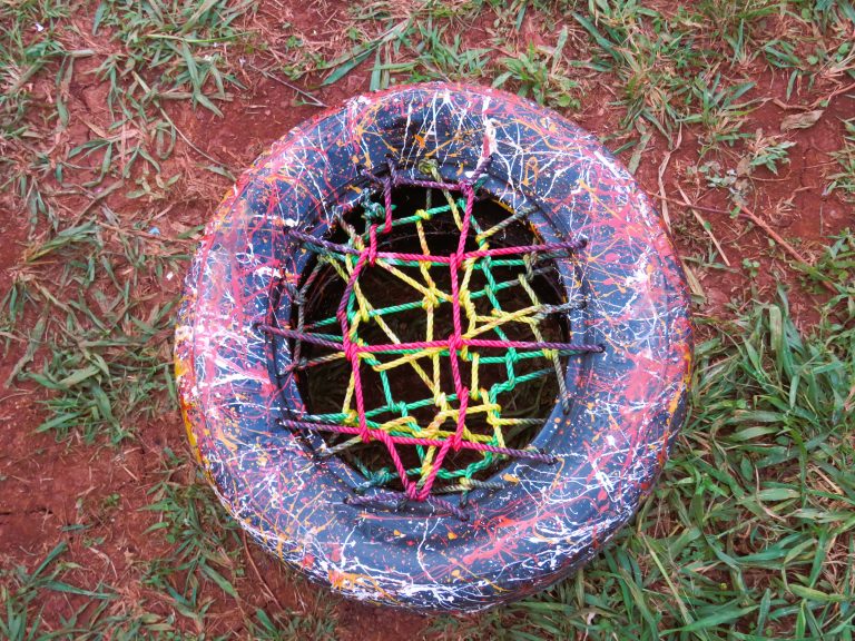 A colorful, hand-painted tire with a net of brightly colored ropes crossed over an open center, resting on a grassy surface