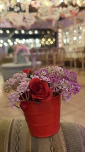 A small red bucket filled with artificial flowers, including red roses and purple blooms, sits in the foreground. The background is softly blurred, revealing a softly lit space with twinkling lights and stylish seating.