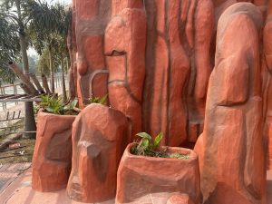 Red rock formation with carved planters containing green plants. Lush palm trees are visible in the background, creating a natural, earthy scene.
