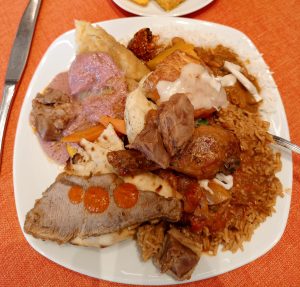 Top-down view of a white plate with roast beef in red sauce, grilled chicken, seasoned rice,  flatbread, and steamed vegetables, served on an orange textured tablecloth.