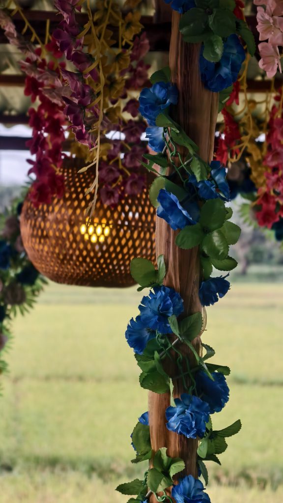 A festive ceiling decorated with colorful flower garlands and a glowing woven bamboo lantern.