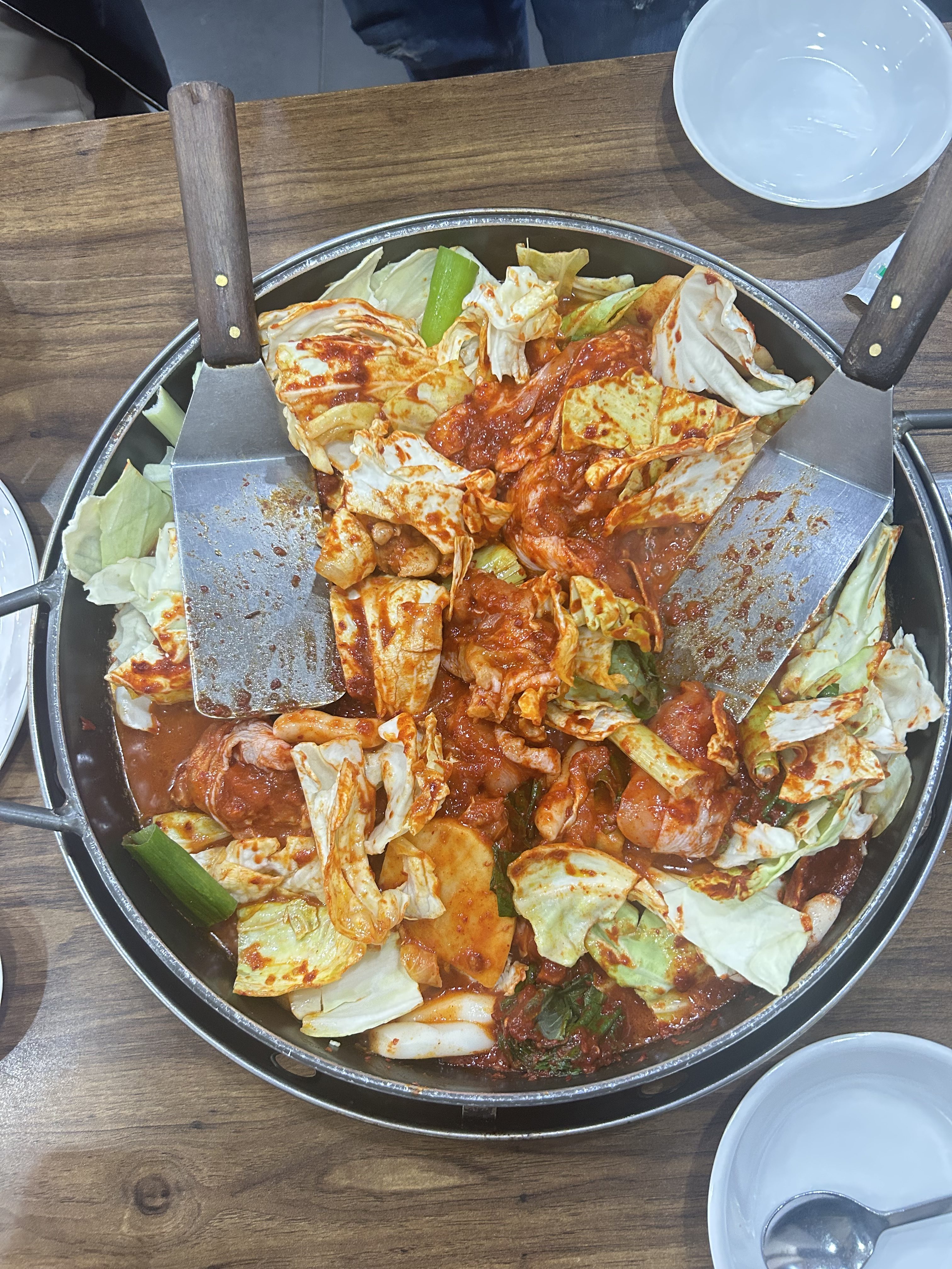 A large, round metal pan sits on a wooden table, filled with a vibrant mix of ingredients. The dish features chunks of cabbage and a variety of sliced vegetables, all coated in a thick, reddish-orange sauce that suggests a spicy flavor.