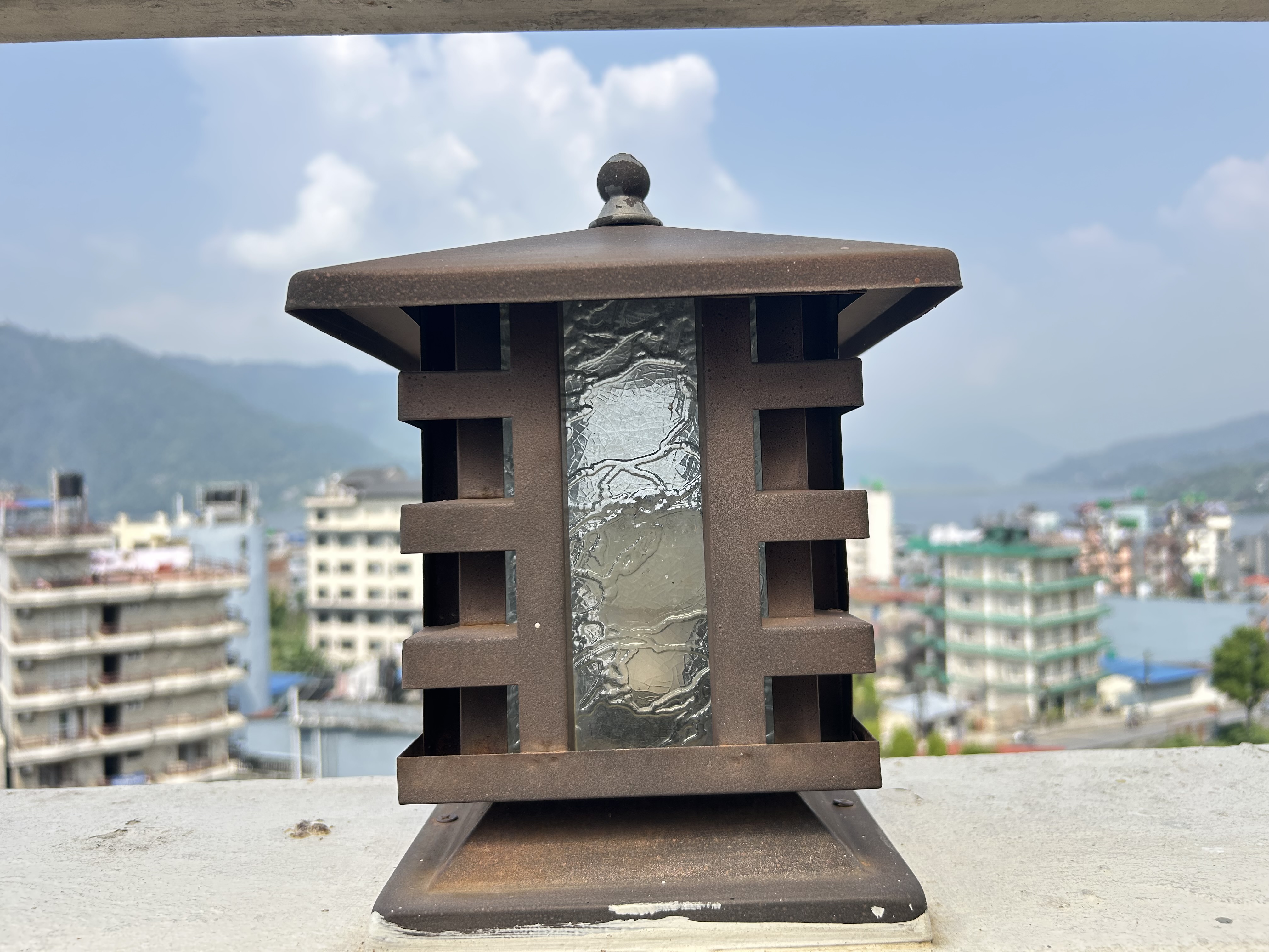 The image features an ornate, square metal lantern with a rustic brown finish. Its design includes vertical and horizontal slats that create a distinctive pattern.
