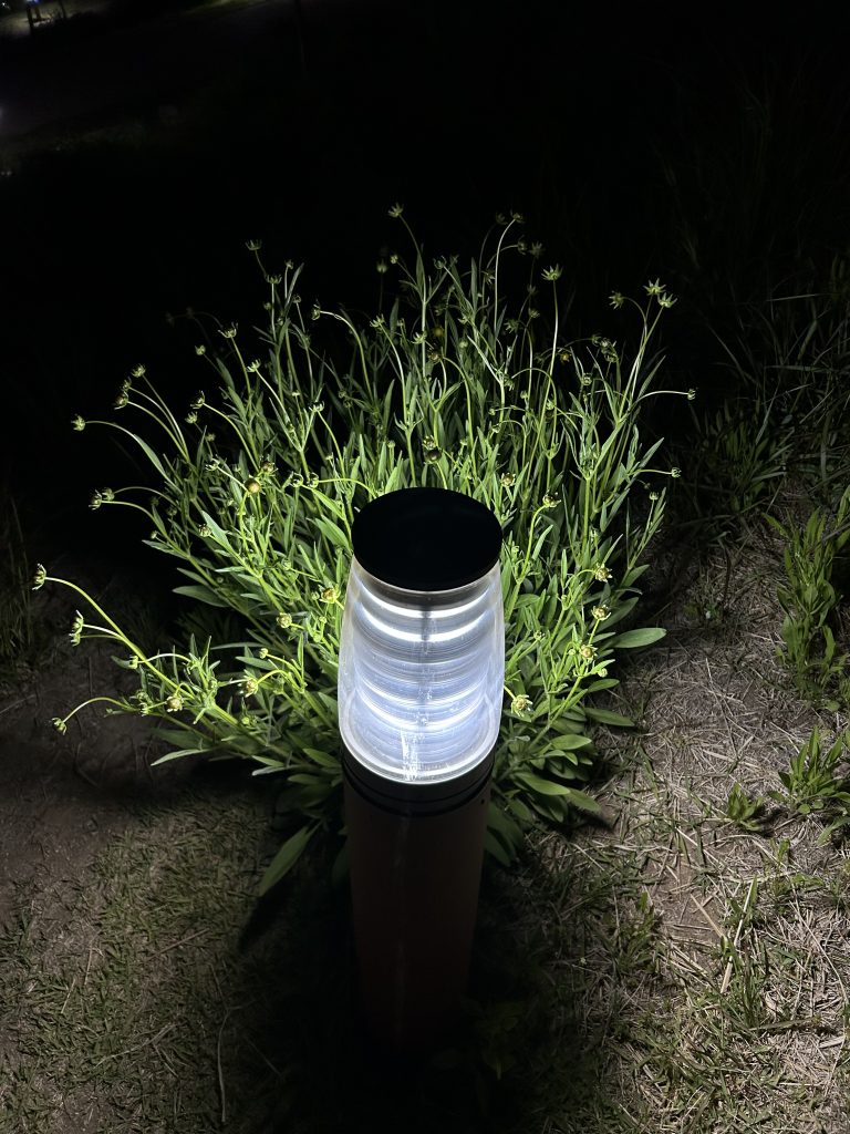 A slim, tall outdoor light stands in a dark setting, glowing brightly from the top with a circular, translucent light source that emits a warm, white light.