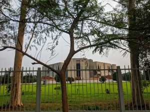The Jatiya Sangsad Bhaban with its geometric design is seen through a metal fence, with green grass and a tree in the foreground, creating a serene atmosphere.