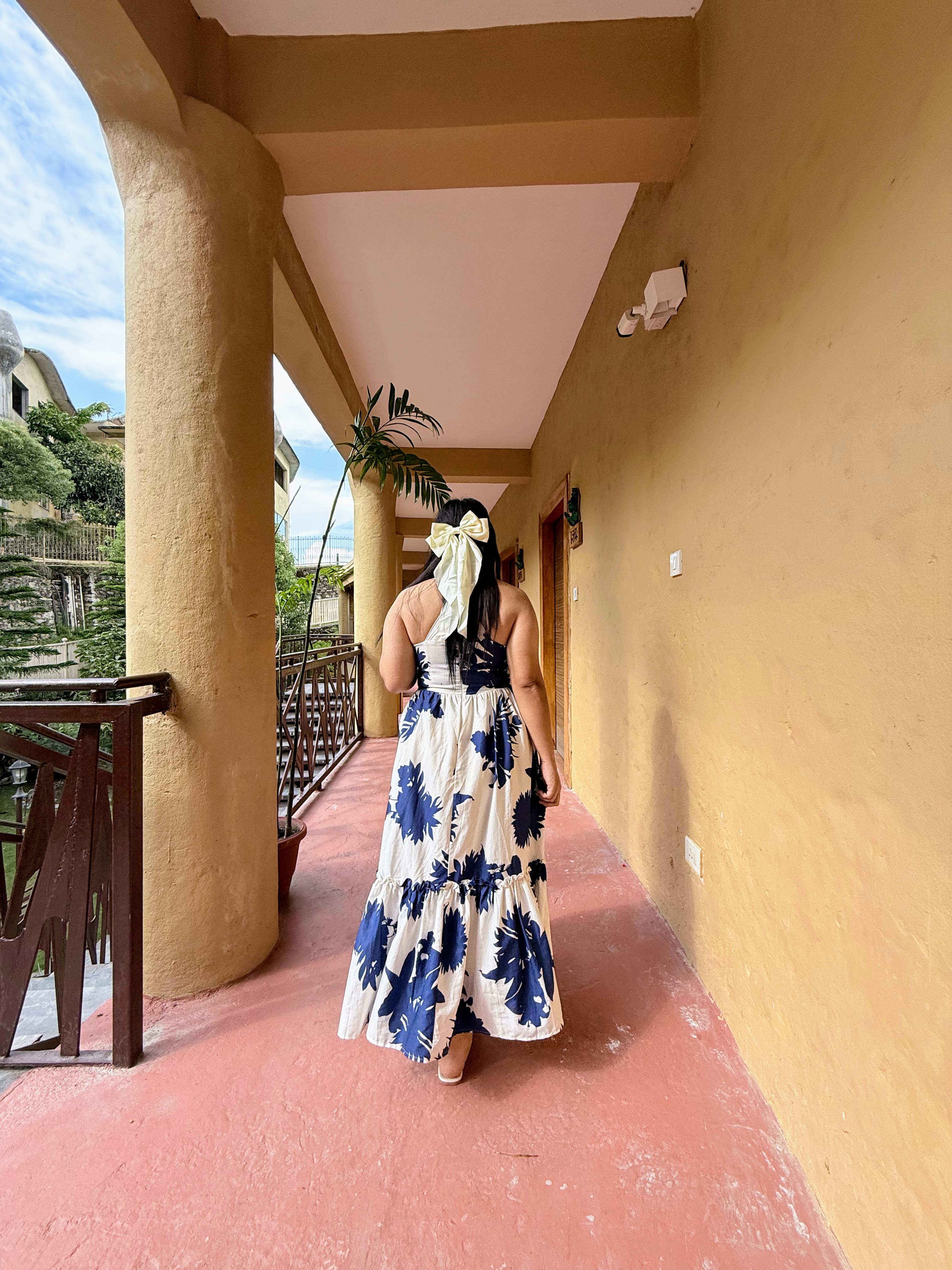 A woman with a large white hair bow walking away down an outdoor building corridor, wearing a white dress with large blue floral patterns.