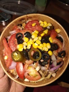 A colorful salad in a brown bowl filled with sliced tomatoes, black olives, corn, red onions, jalapeños, and grilled chicken, creating a fresh, vibrant look.