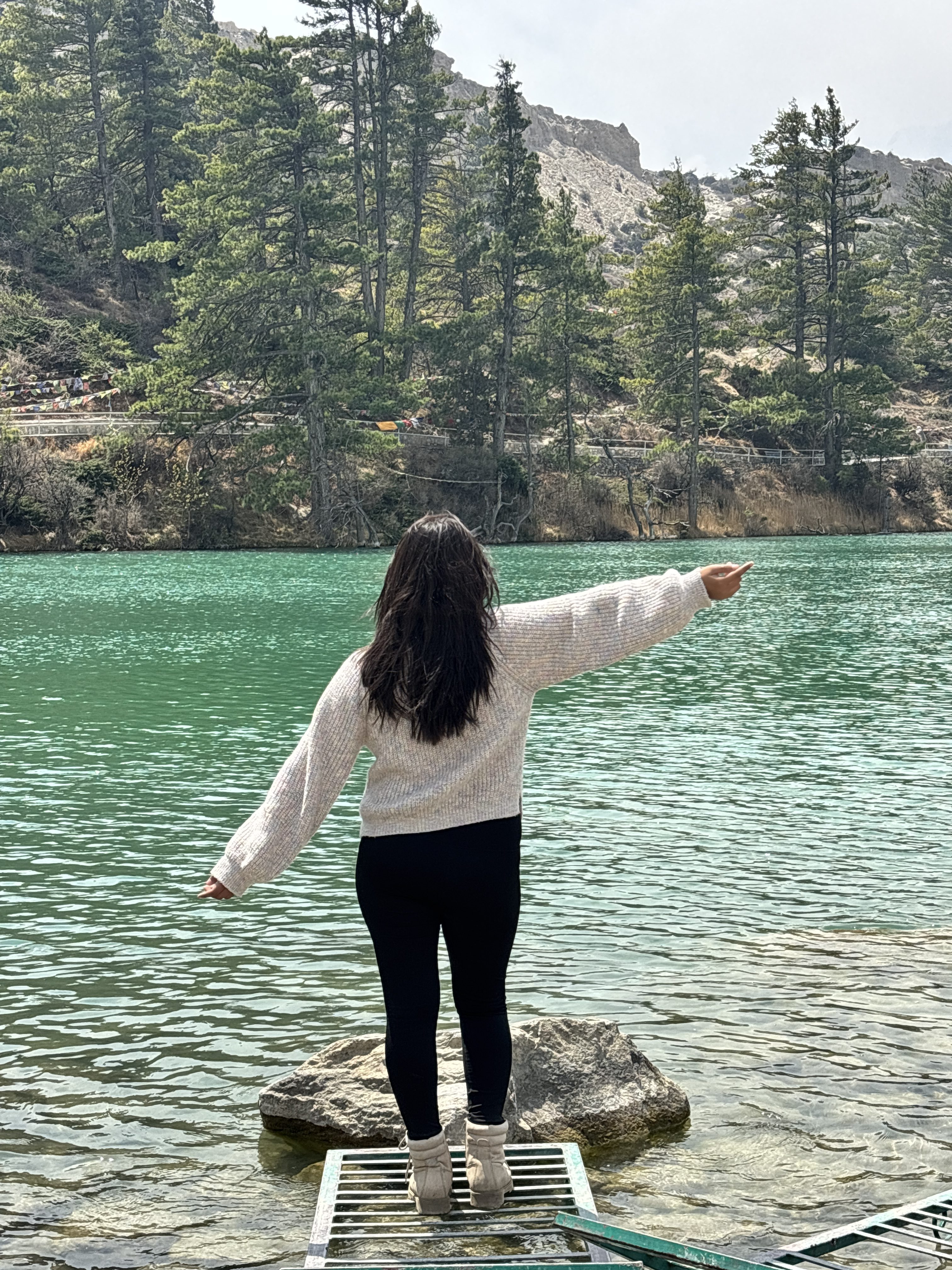 Woman in a white sweater stands on a rock, arms outstretched, facing a serene turquoise lake surrounded by pine trees and rocky hills.