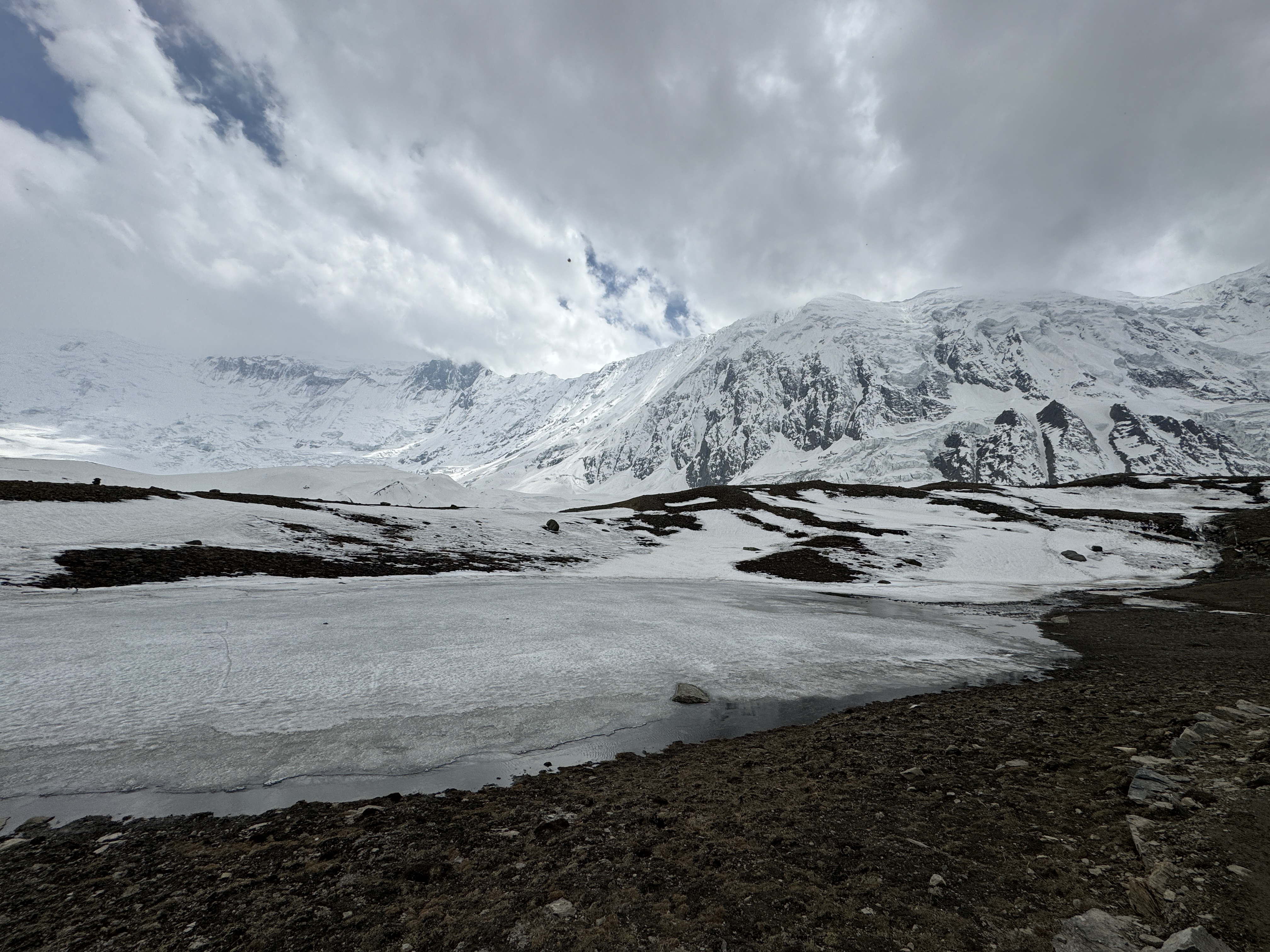 A partially frozen glacial lake in the foreground with a massive, snow-covered mountain ridge rising behind it, captured near Tilicho Lake at an elevation of 4,919 meters in Nepal.