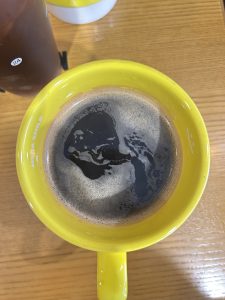 Yellow mug filled with black coffee sits on a wooden table. The coffee's surface has a swirling pattern. A sense of warmth and relaxation is conveyed.

