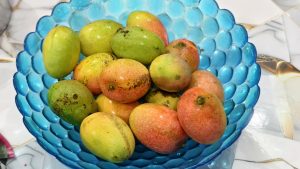 A top-down view of a blue glass bowl filled with many small mangoes. The mangoes are in various stages of ripeness, displaying shades of green, yellow, and bright orange-red. The bowl sits on a white tiled surface with a subtle marble pattern.