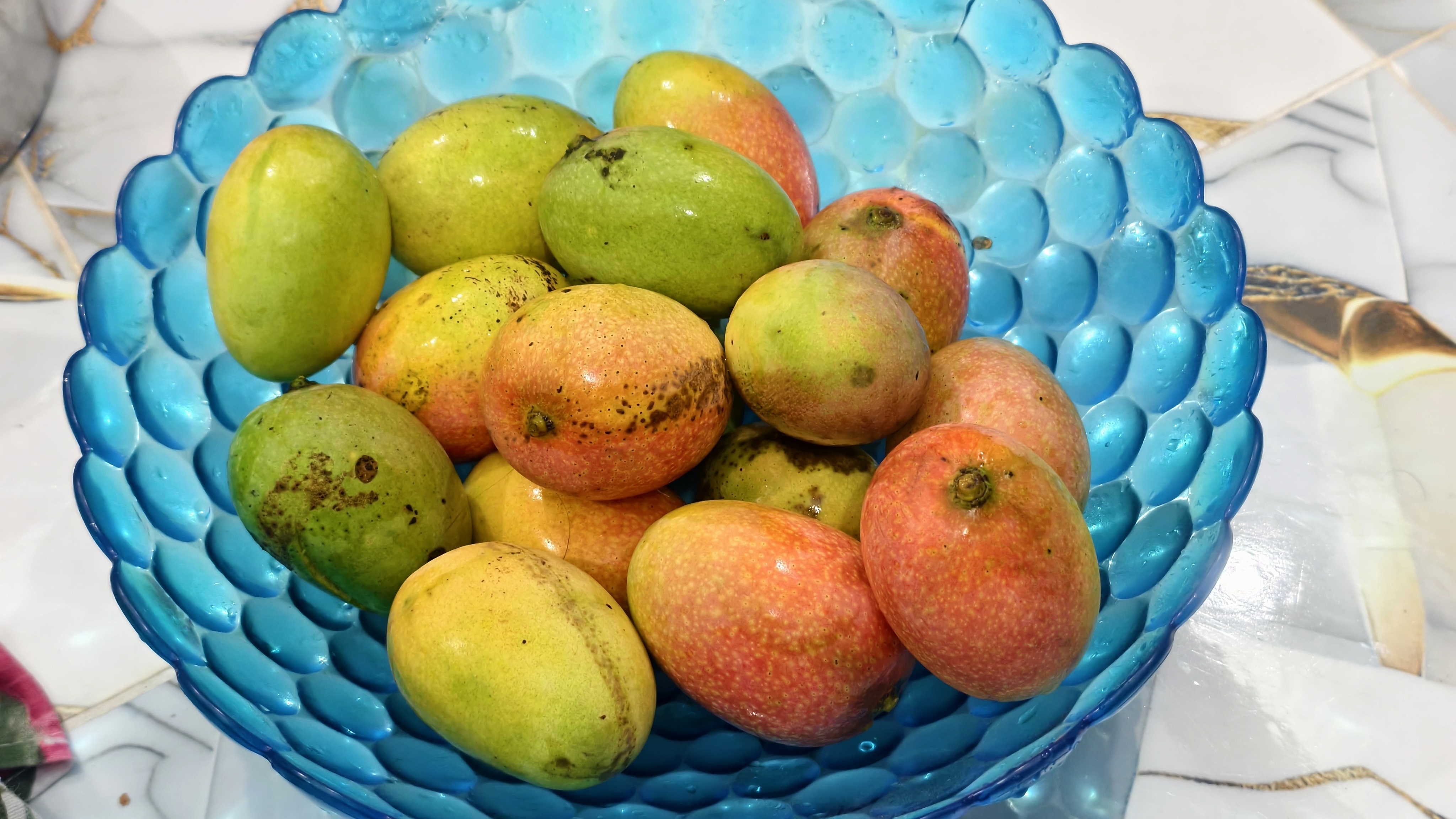 A top-down view of a blue glass bowl filled with many small mangoes. The mangoes are in various stages of ripeness, displaying shades of green, yellow, and bright orange-red. The bowl sits on a white tiled surface with a subtle marble pattern.