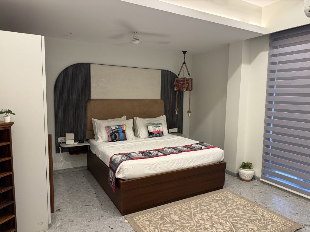 A cozy bedroom with a neatly made bed, colorful pillows, a textured headboard, bedside lamp, and a modern wooden bookshelf.