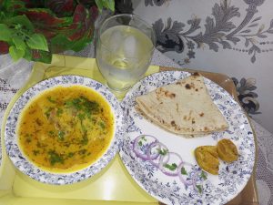 A Pakistani meal featuring dal in a floral bowl, chapati, sliced onions, lemon pickle on a plate, and a glass of ice water on a yellow tray.
