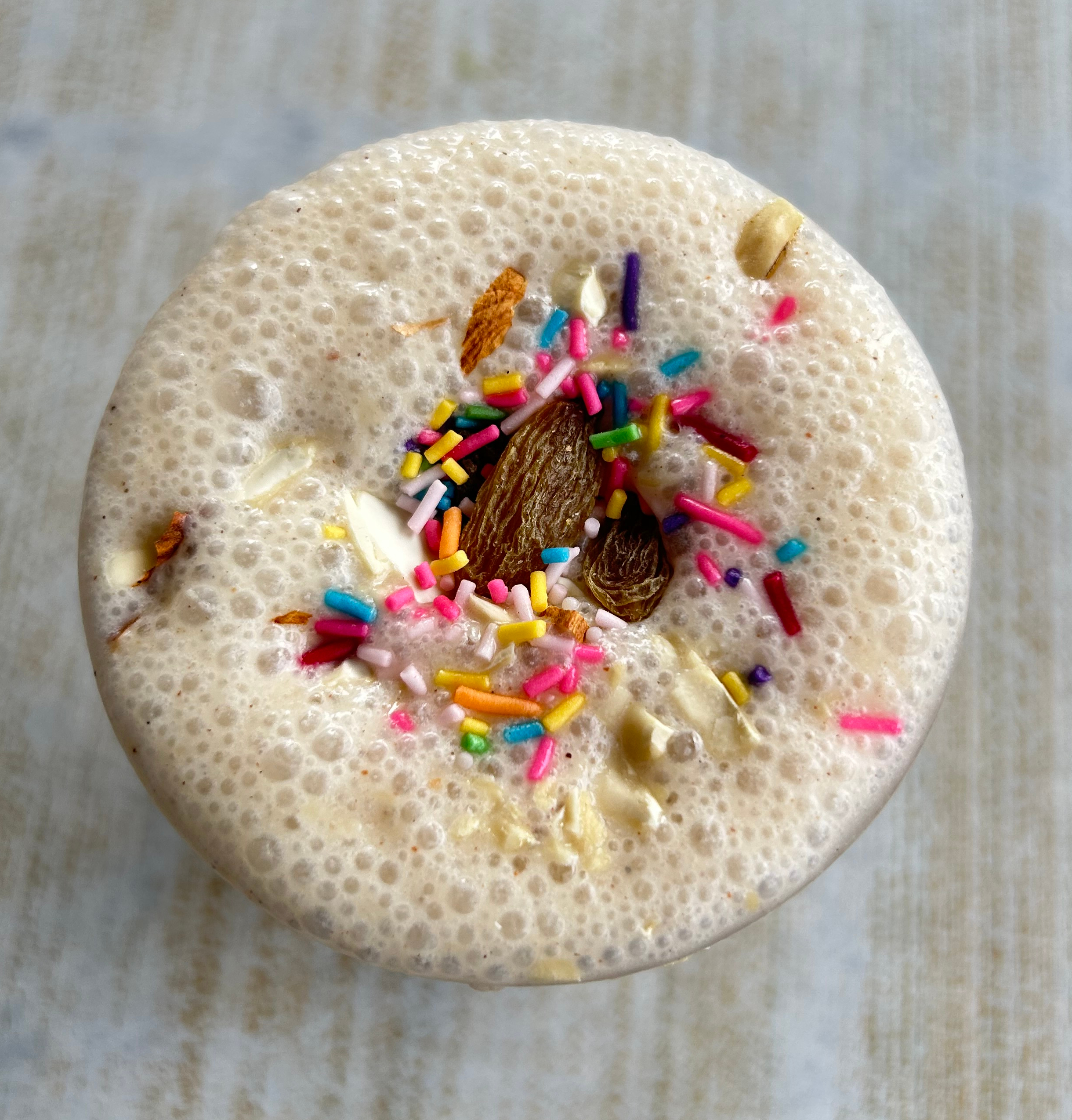 A frothy beverage topped with colorful sprinkles and almonds sits on a light wooden table.