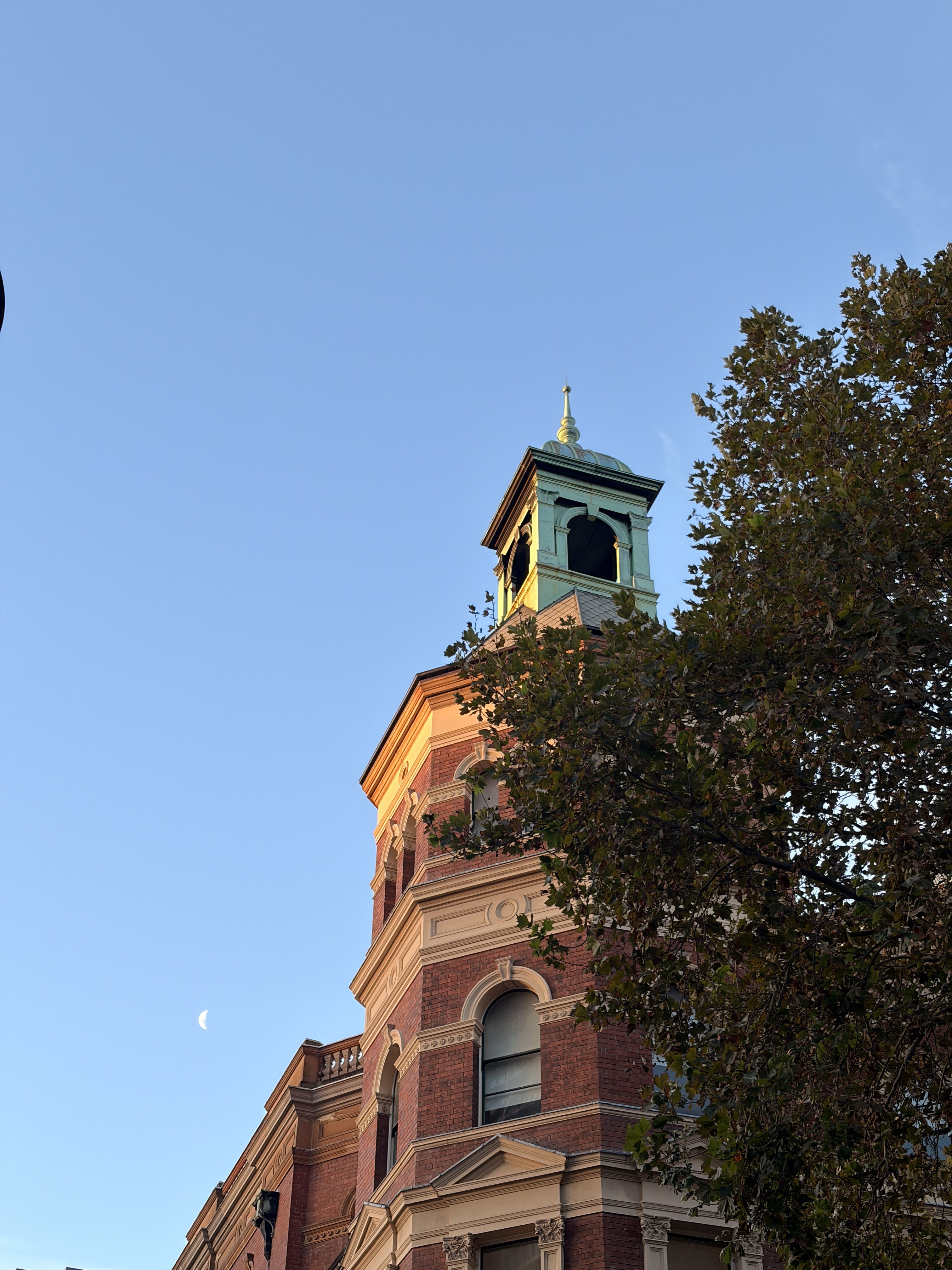 Historic brick building with a green-capped tower against a clear blue sky. The moon is visible to the left, and trees partially frame the scene.