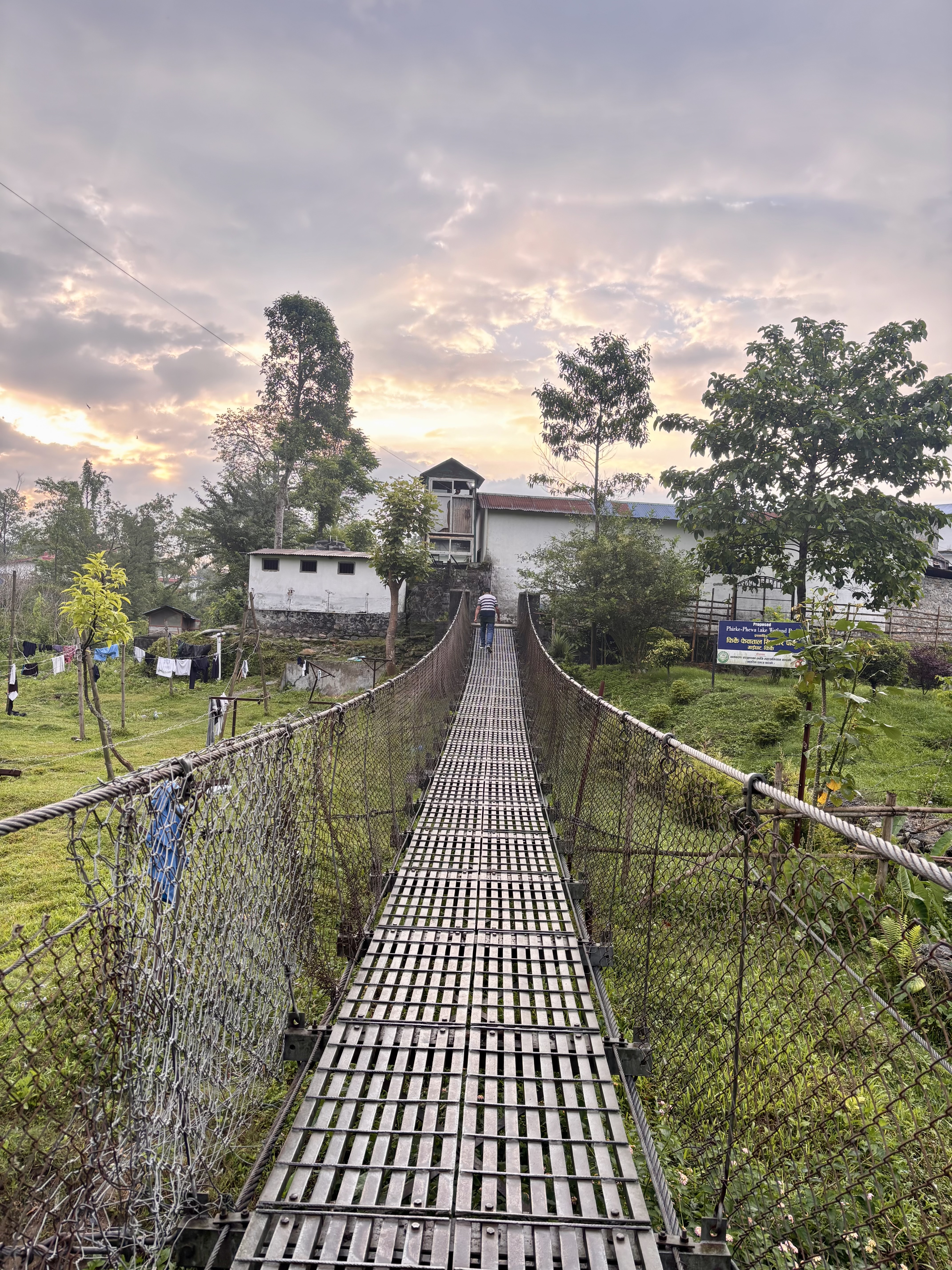 Suspension bridge leading to a white house, surrounded by lush greenery under a colorful sunset sky, with a person walking across.