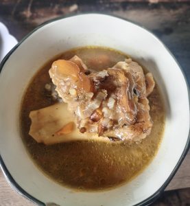 A white bowl of clear broth with a piece of cooked cow’s foot (molokoni) and vegetables.