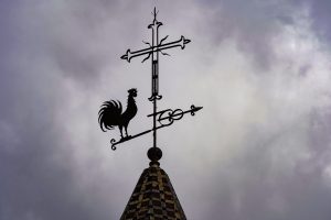 A decorative weather vane sits atop a tall, pointed rooftop. The silhouette of a rooster is prominently featured, standing on a horizontal arm that extends outward. Above the rooster, there is a cross-shaped design, adorned with ornate details. 
