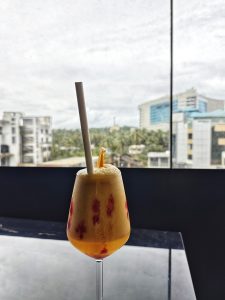 A colorful juice in a tall glass on a dark surface, with a cloudy sky and buildings visible through a window in the background.