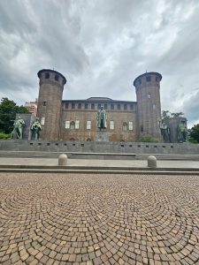 A grand historic castle (Monumento a Emanuele Filiberto Duca d'Aosta) with tall towers and statues stands under dramatic cloudy skies. The wide stone courtyard adds depth and symmetry. Captured in Turin (Torino), Italy.