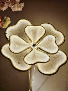 A flower-shaped decorative light with a soft golden glow stands against a warm wall, with artificial flowers beside it. Shot indoors in Perumanna, Kozhikode. 