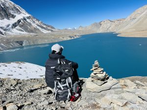 A person in outdoor gear sits beside a cairn overlooking turquoise Tilicho Lake, surrounded by snow-capped mountains under a clear blue sky.