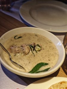 A white bowl of creamy, beige soup with herbs and chicken pieces. A spoon rests inside. 